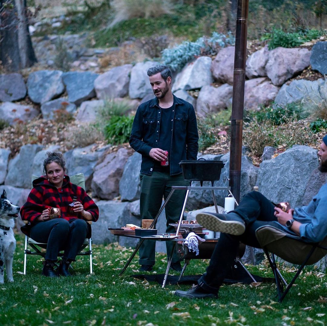 Winnerwell® Fire Guard Stove Table as part of a group camping setup, with people gathered around, illustrating its role in social outdoor cooking activities.