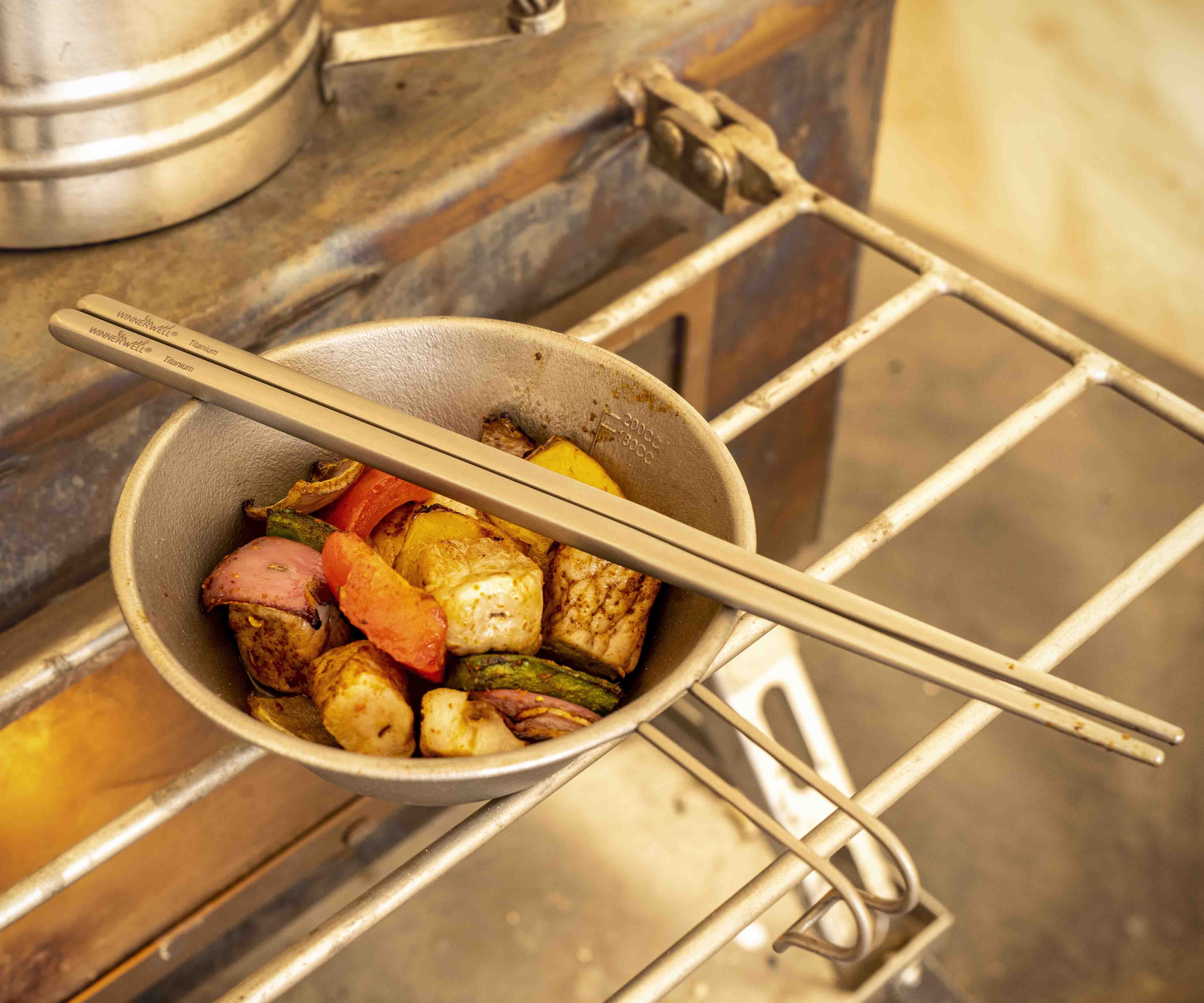Winnerwell® square titanium chopsticks being used to cook a mix of ingredients on a grill, showcasing their use in outdoor culinary activities.