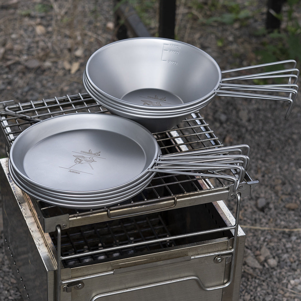 Winnerwell® stainless steel Sierra cooking bowls and plates placed on an outdoor grill, ready for campfire cooking.