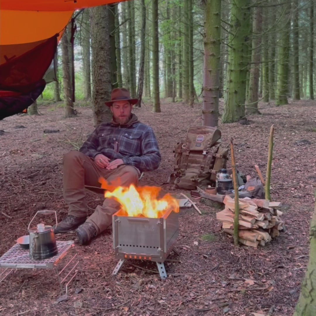 Winnerwell Secondary Combustion Portable Grill Firepit demonstrated in a forest campsite, showing how it creates a cozy fire and serves as a cooking station for outdoor enthusiasts.