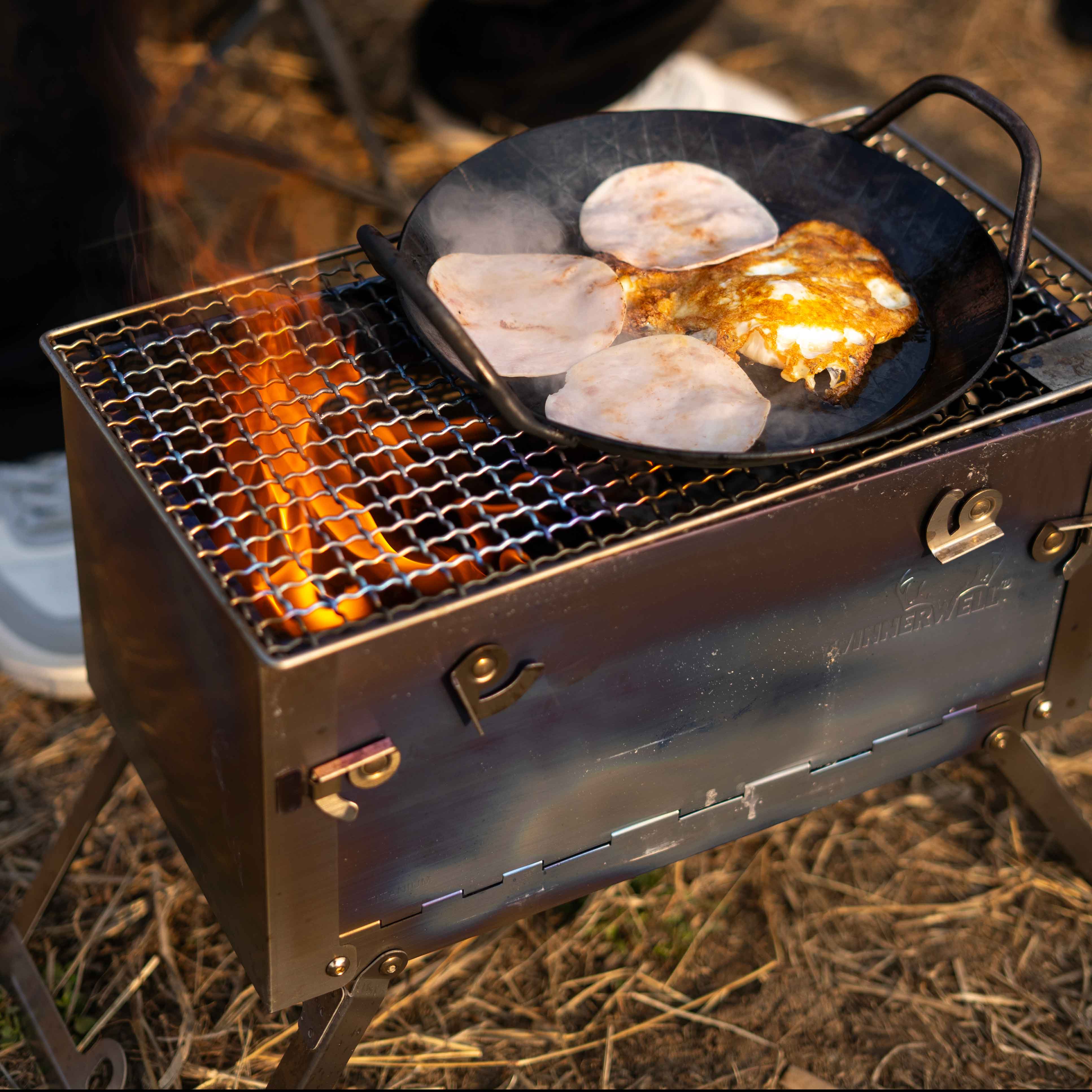 Winnerwell® Fastfold Titanium Camping Stove Series in use, cooking in the wild. Made of titanium, it provides efficient heat for outdoor cooking, ideal for preparing meals during camping adventures, showcasing its practical cooking function.