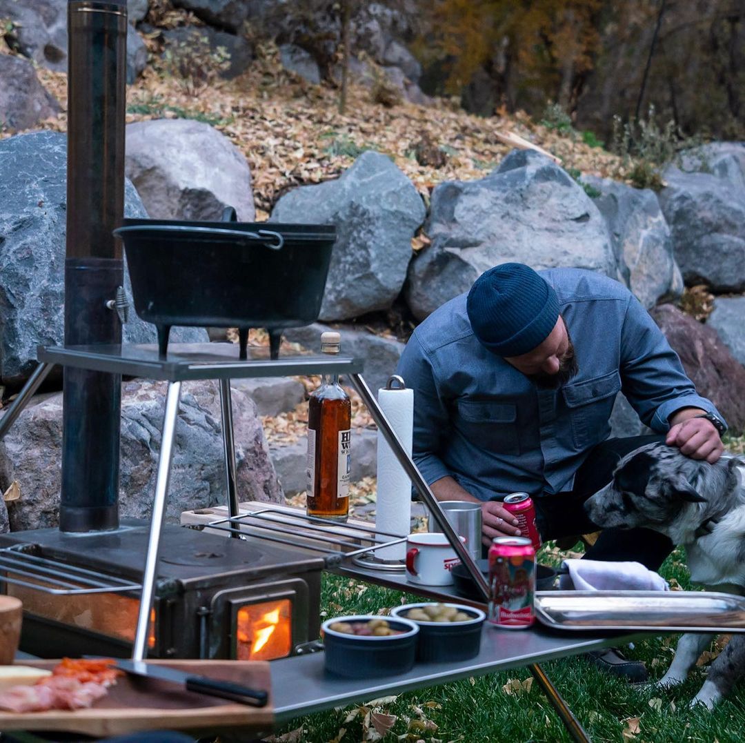 Winnerwell® Fire Guard Stove Table being used in a natural outdoor setting, with a person cooking over a fire beneath it, demonstrating practical camp - cooking application.