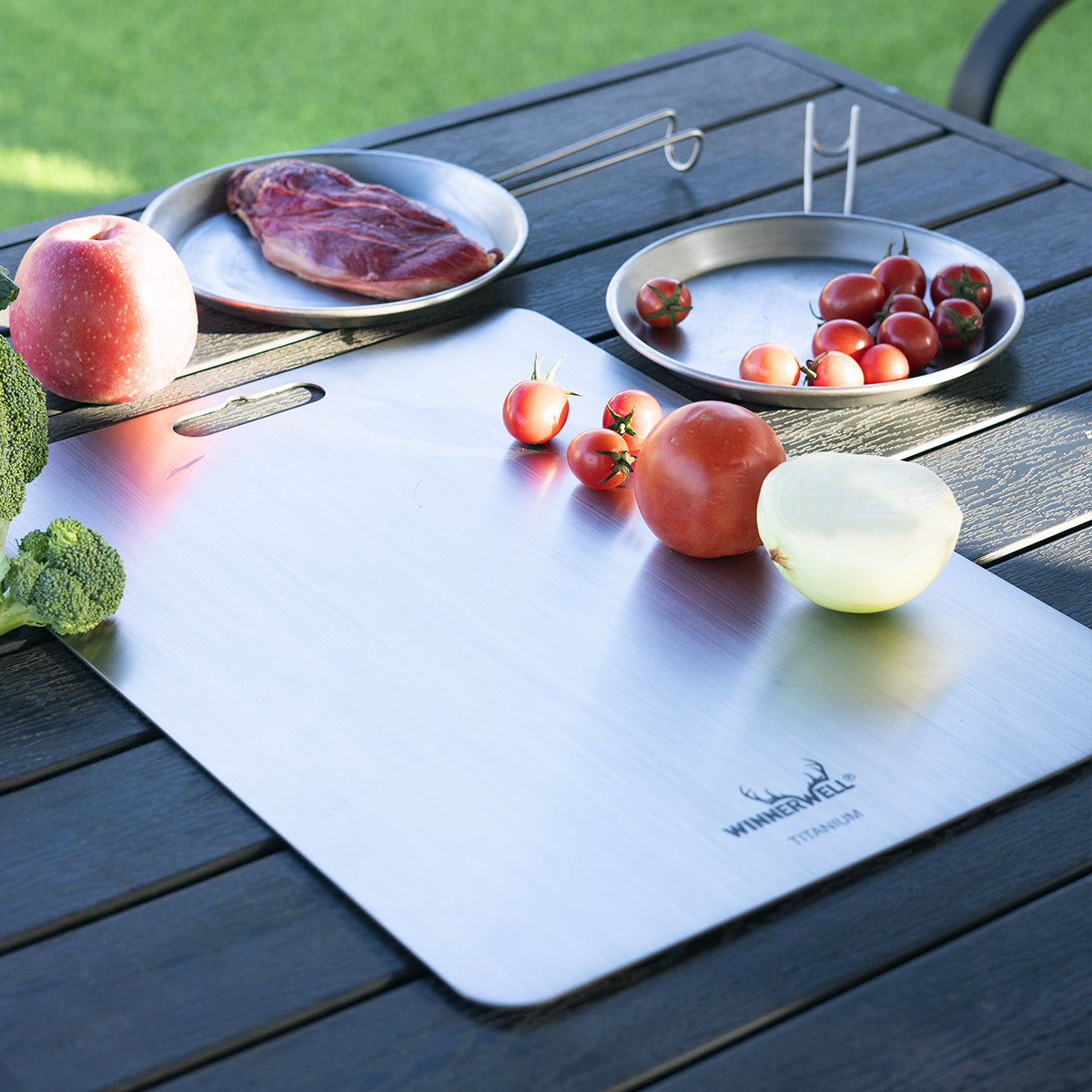 Winnerwell® titanium cutting board placed on an outdoor table with fruits, demonstrating its use in a picnic or outdoor cooking scenario.