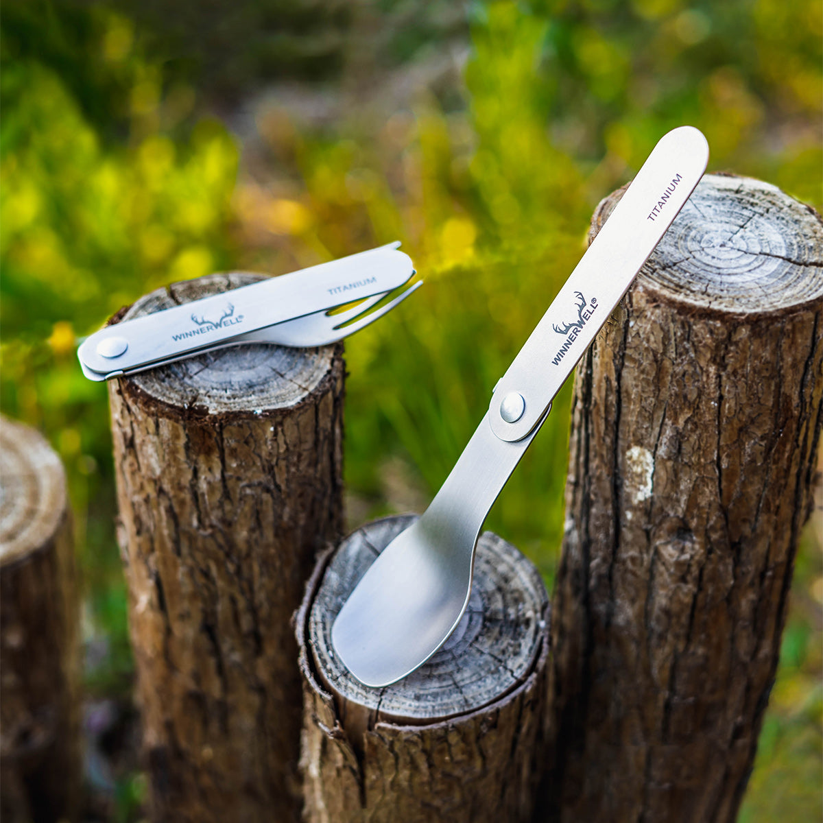 Winnerwell® titanium folding fork and spoon placed on wooden stumps in a natural outdoor environment, highlighting their portability for camping or hiking.