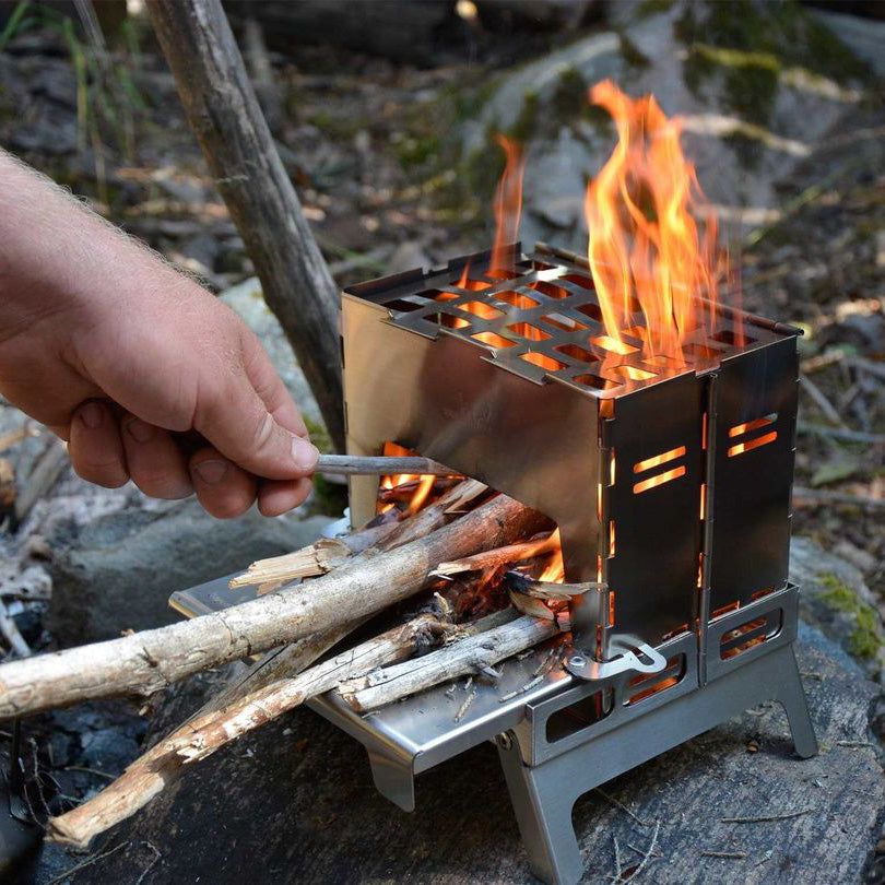 Winnerwell® Backpack Stove in outdoor use, featuring a wood - burning design with visible flames. It's a foldable, portable camping stove. Ideal for outdoor camping, picnics, and wild cooking, made of durable stainless steel/titanium, easy to set up and use for a convenient outdoor cooking experience. 