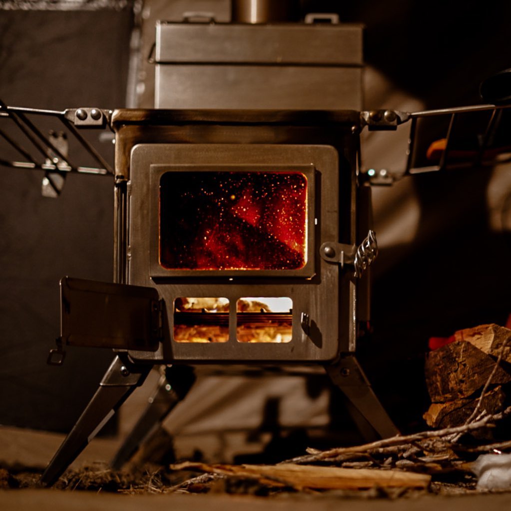Winnerwell® Nomad View 1G Cook Camping Stove Package in outdoor burning setup. Shows the stove with a visible fire inside, arranged in a campsite environment. The foldable, stainless - steel construction is designed for reliable outdoor cooking, heating, and easy transport—ideal for wilderness adventures and group camping trips.